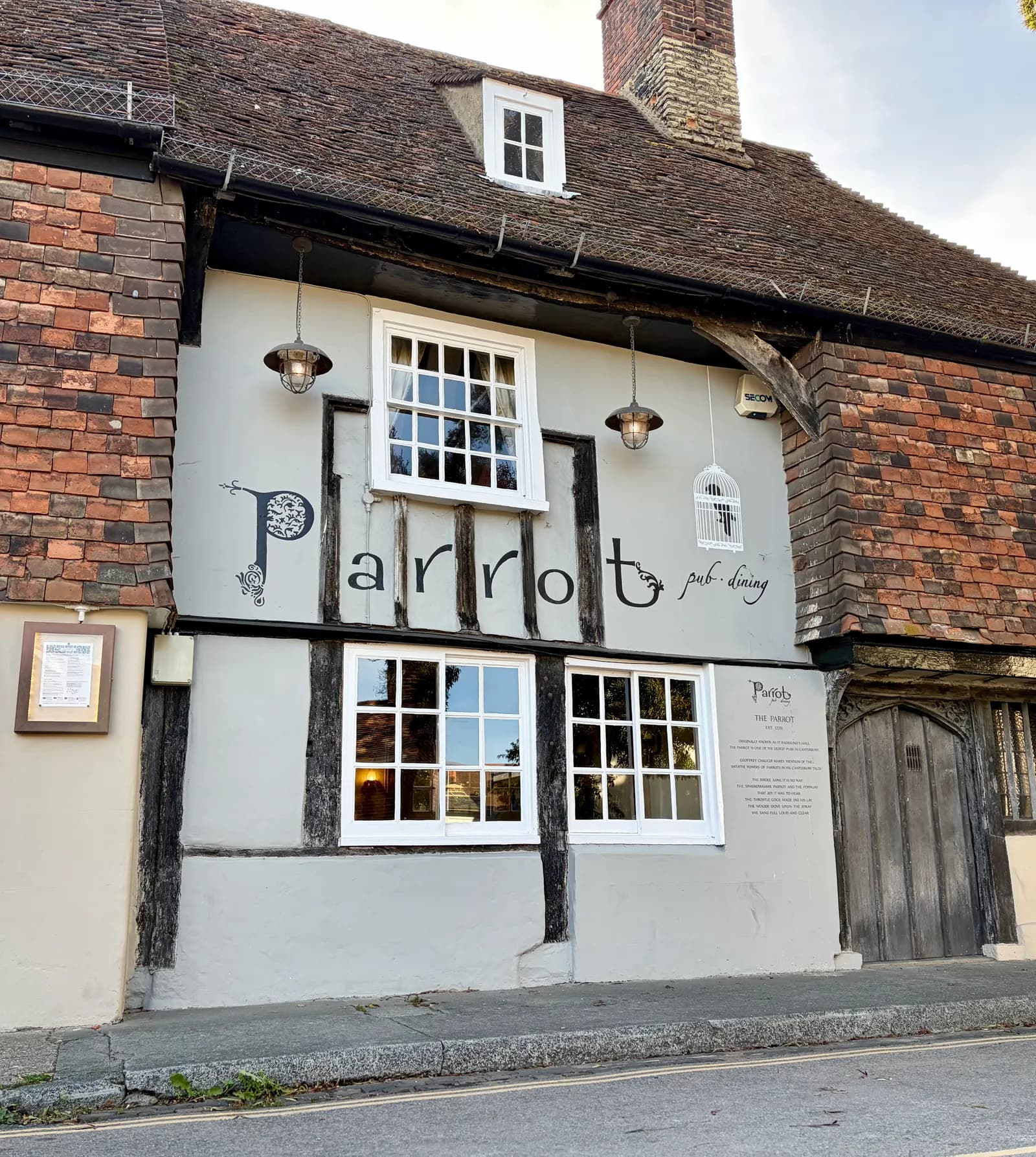 The exterior of The Parrot pub and dining establishment, featuring white windows, decorative hanging lamps, and black text against a light gray facade, all beneath a rustic tiled roof.